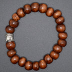 A beaded bracelet made of glossy medium-brown wooden beads with a small silver Buddha head charm, displayed on a neutral gray background.