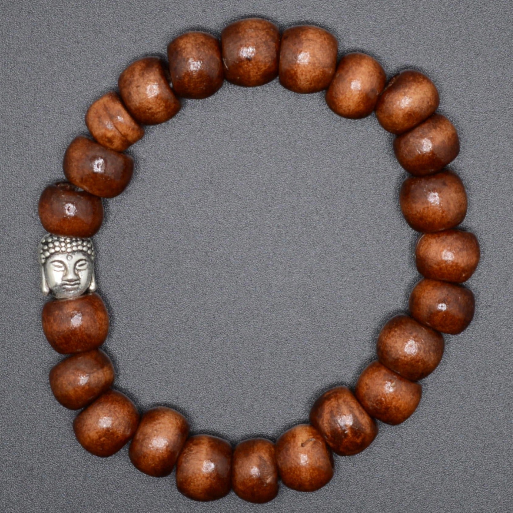 A beaded bracelet made of glossy medium-brown wooden beads with a small silver Buddha head charm, displayed on a neutral gray background.