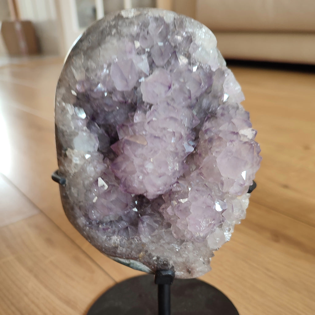 A large amethyst crystal geode cluster on a black display stand, photographed indoors on a wooden floor with a blurred living room background.
