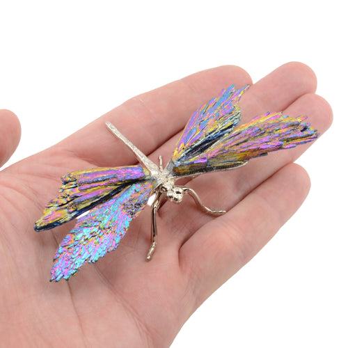 A metallic dragonfly wing ornament with iridescent purple, blue, and gold foil details, resting on a person's palm against a white background.