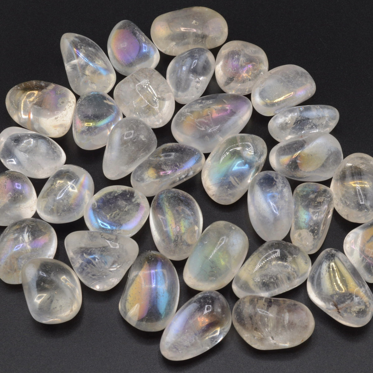A pile of polished tumbled aura quartz stones with an iridescent rainbow sheen, arranged on a dark black background.