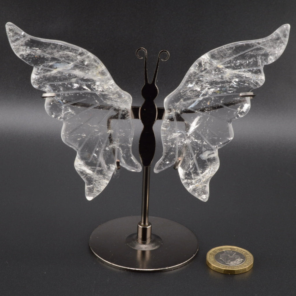 A clear quartz crystal butterfly wing holder with a black metal body and stand, displayed on a dark gray background with a Euro coin for scale.