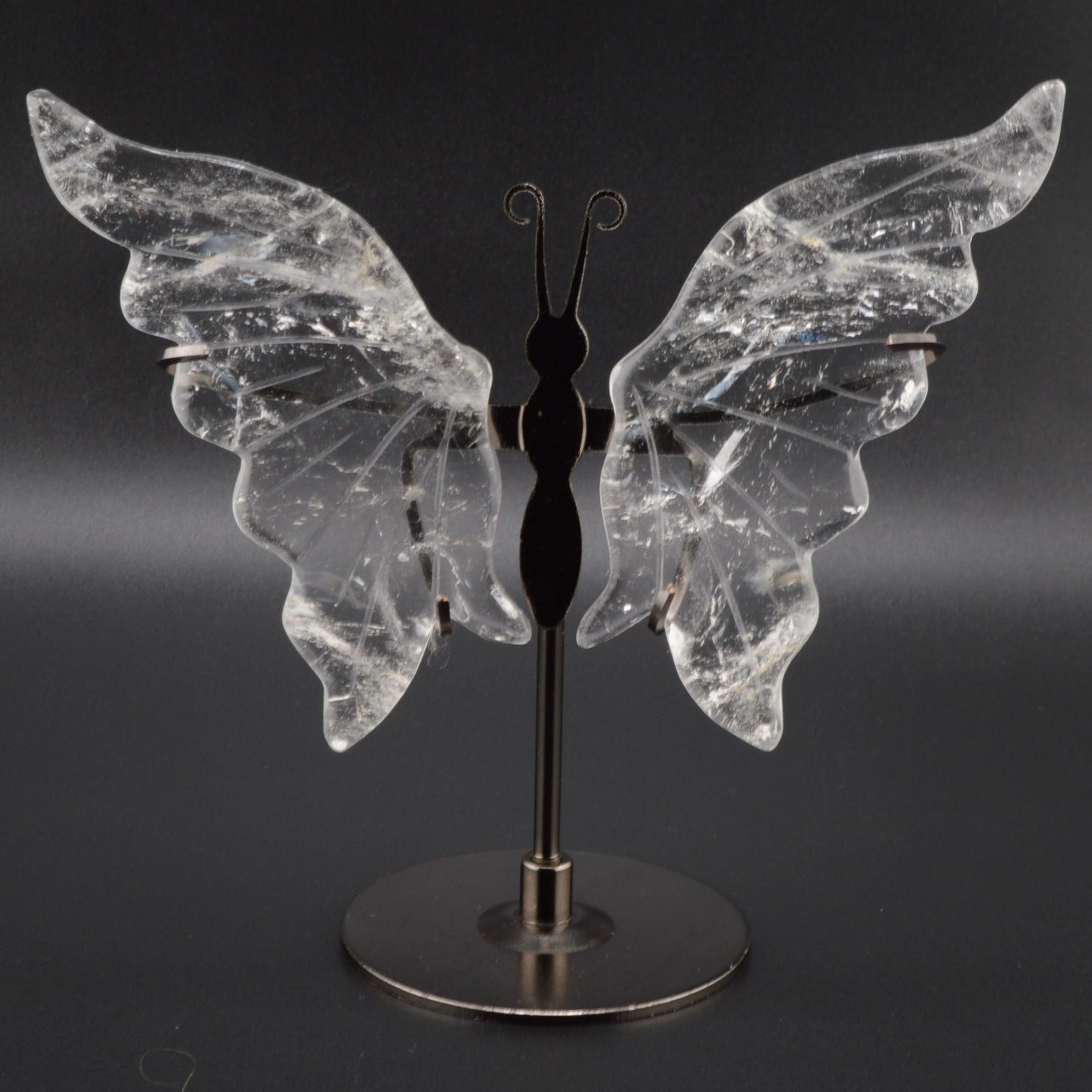 A butterfly-shaped crystal sculpture with clear quartz, inclusions-filled wings mounted on a black metal stand with a round base, photographed against a dark gray background.