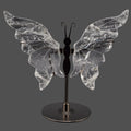 A decorative butterfly-shaped shelf piece with clear quartz crystal wings mounted on a black metal stand with a round base, shown against a dark gray background.