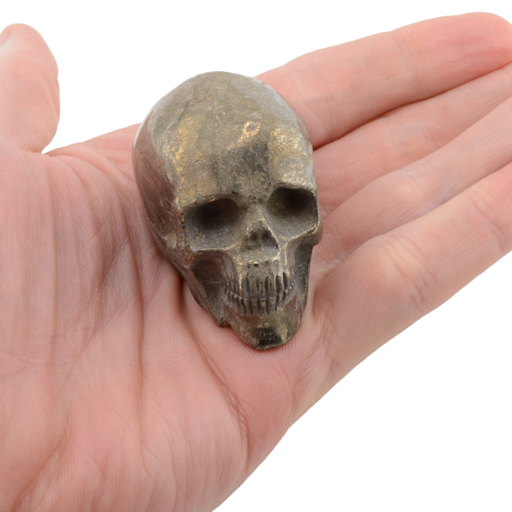 A small metallic pyrite (AKA fools gold) skull figurine with a distressed bronze finish, held in the palm of a hand against a plain white background.
