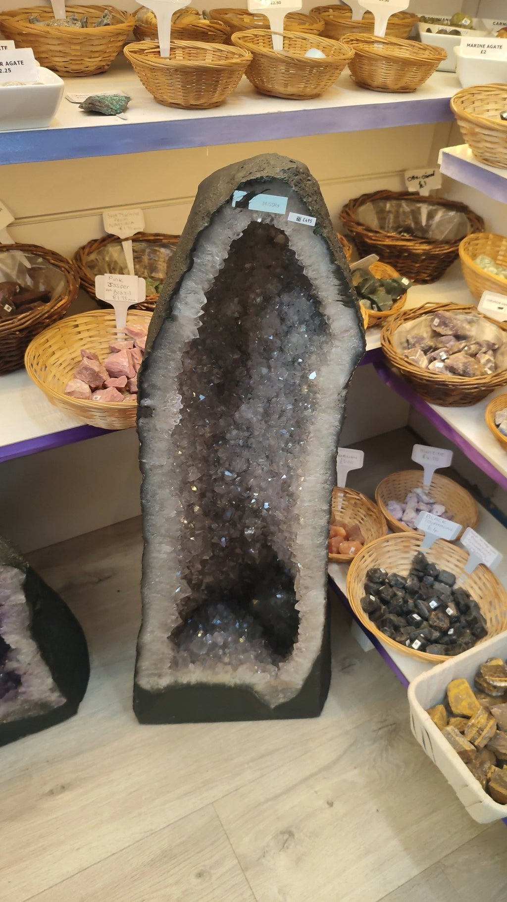 A large natural amethyst geode with a dark gray outer shell and a sparkling purple-gray crystal cavity, displayed on a store shelf surrounded by other stones and baskets.