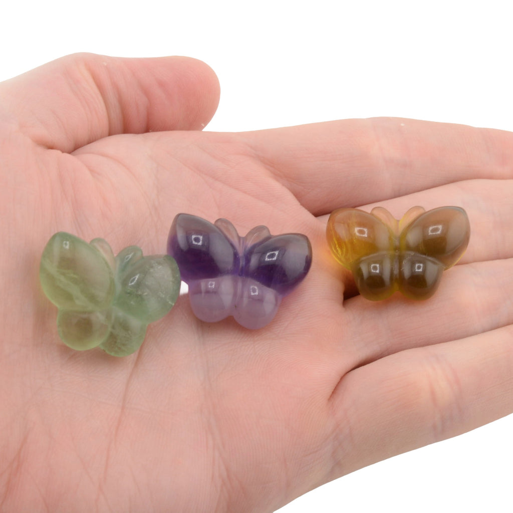 Three small fluorite crystal carved butterflys in green, purple, and amber-brown translucent stone, displayed on an open hand against a white background.