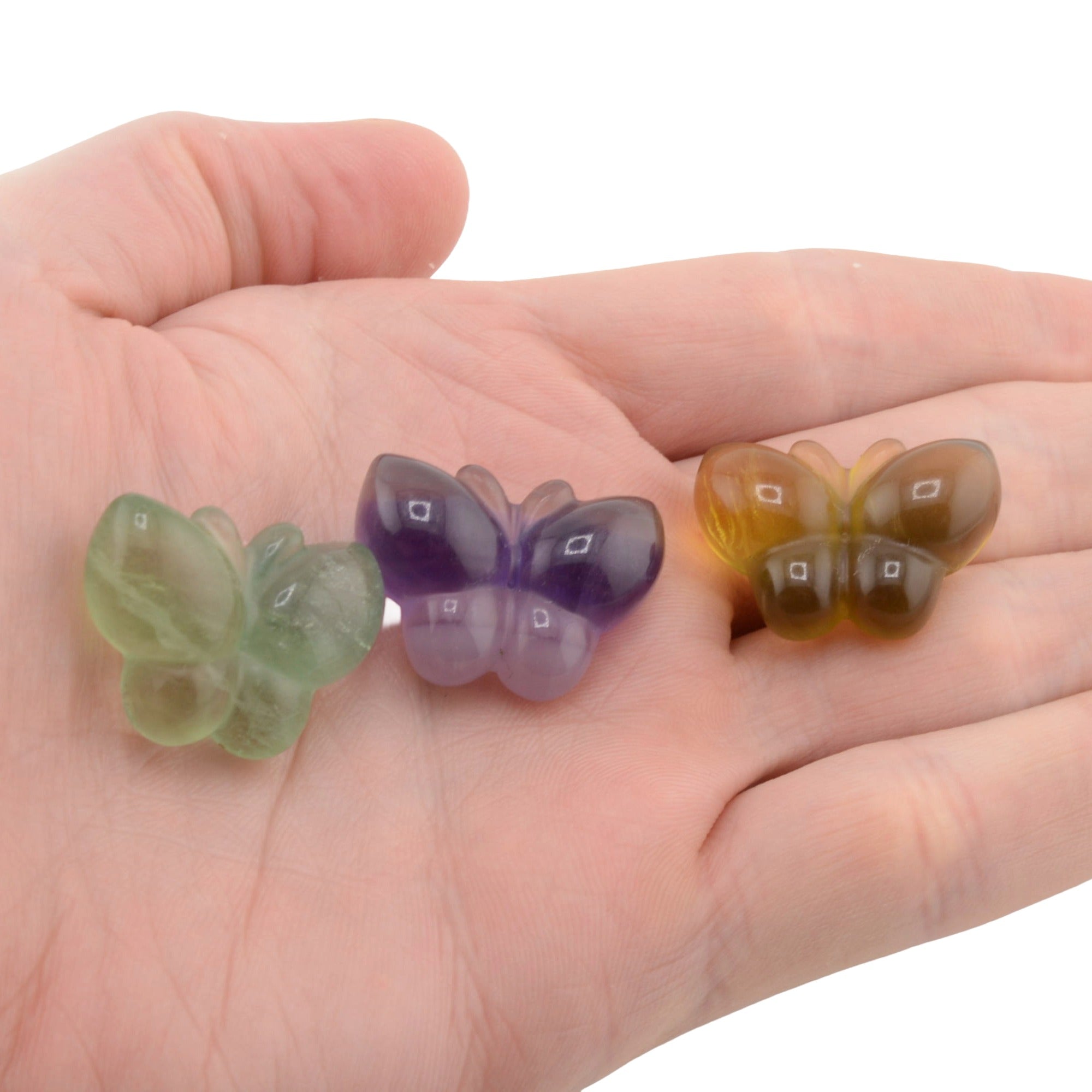 Three small fluorite crystal carved butterflys in green, purple, and amber-brown translucent stone, displayed on an open hand against a white background.