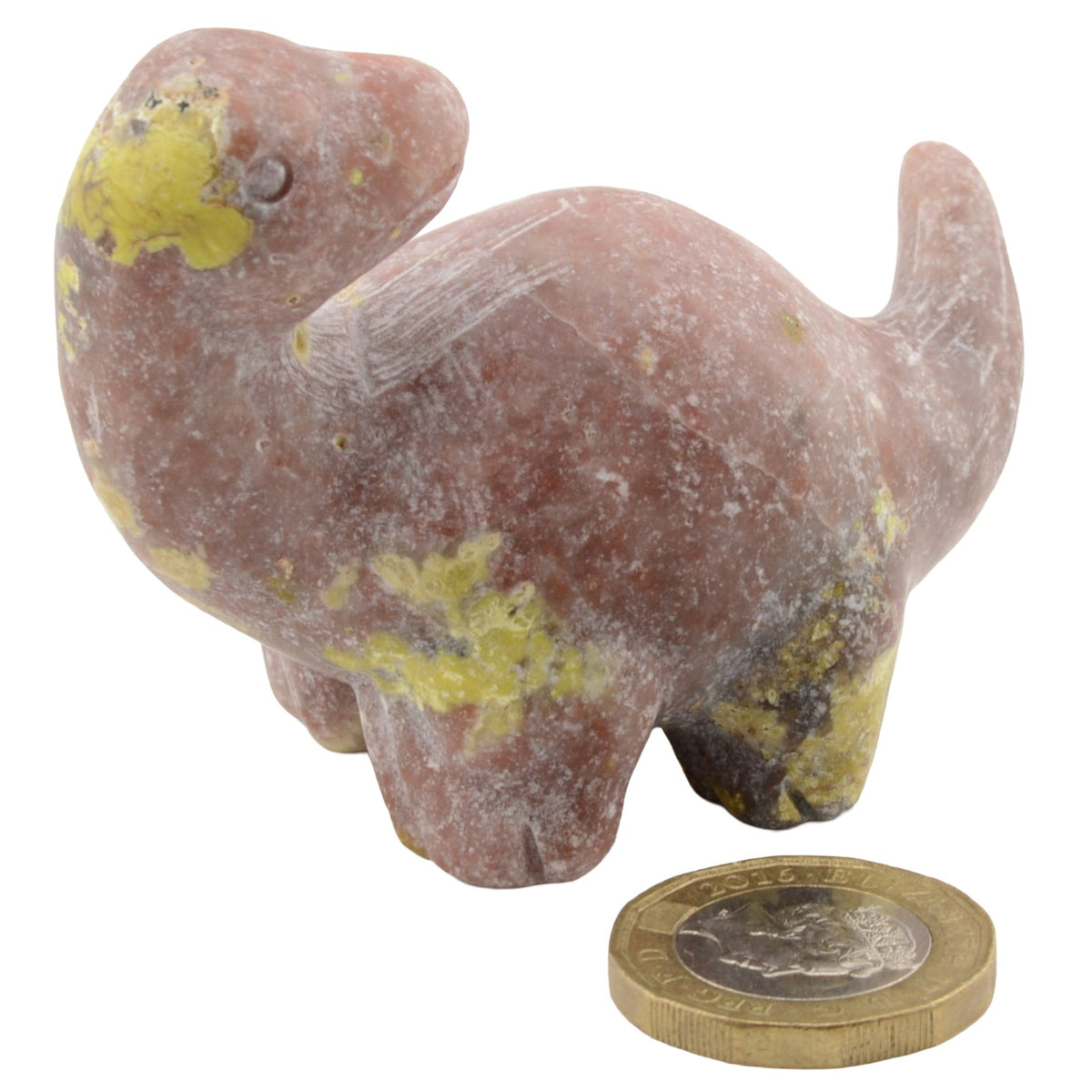 A small carved figurine of a dinosaur made from pinkish gemstone with yellow patina, shown next to a British coin for scale on a white background.