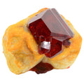 A natural pruskite crystal gemstone specimen featuring a large, faceted deep red crystal mounted on a textured, orange-yellow matrix, isolated on a white background.