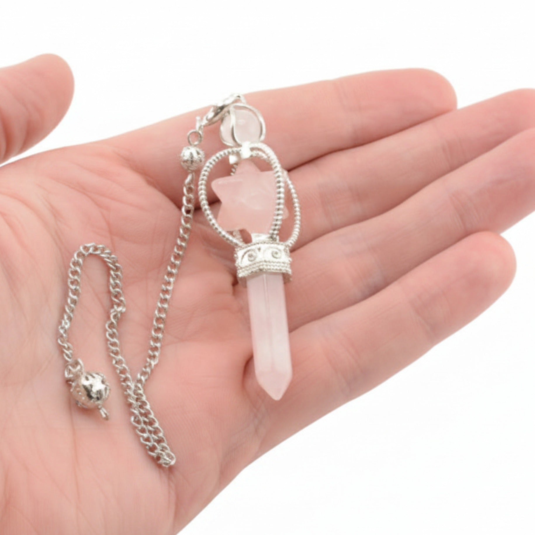 A silver-toned pendulum with a pink rose quartz crystal point and merkaba, displayed resting on an open hand against a white background.