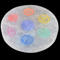 A circular white selenite charging plate featuring seven colourful chakra symbols in orange, yellow, green, blue, and pink, set against a solid black background.
