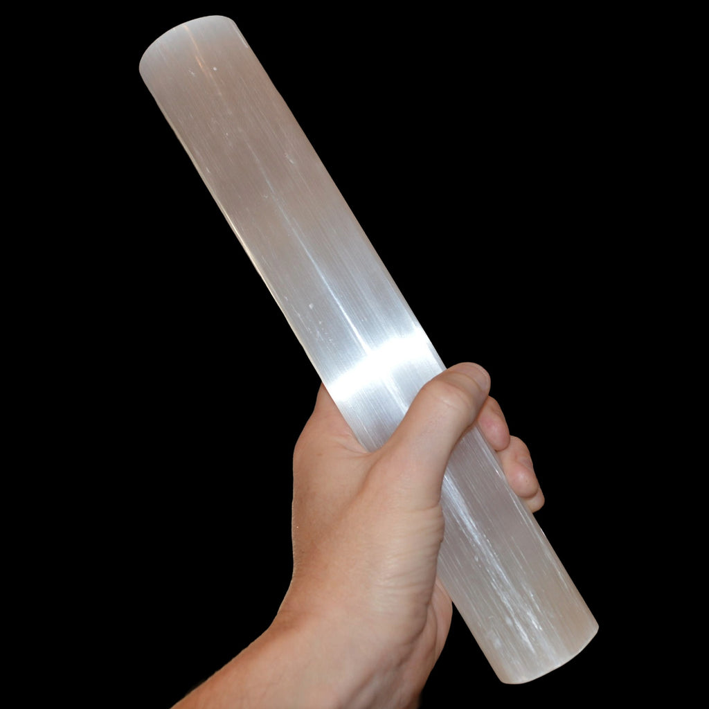 A hand holding a very large, polished white selenite wand against a solid black background.