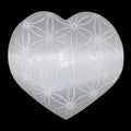 A heart-shaped translucent selenite carving with a white engraving of the flower of life, shown against a solid black background.