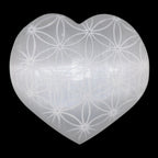 A heart-shaped translucent selenite carving with a white engraving of the flower of life, shown against a solid black background.