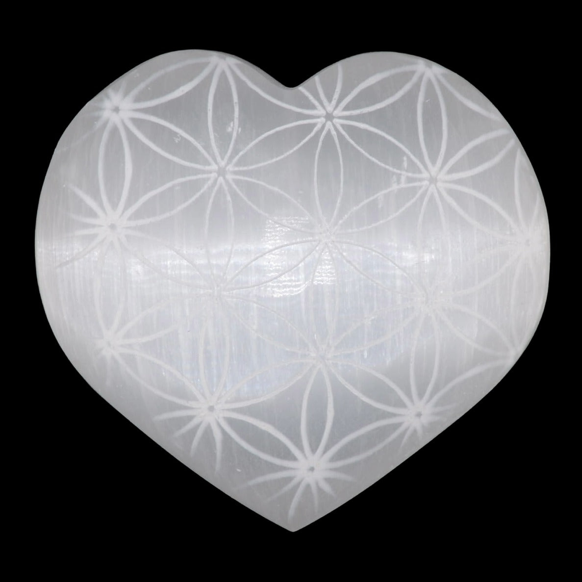 A heart-shaped translucent selenite carving with a white engraving of the flower of life, shown against a solid black background.
