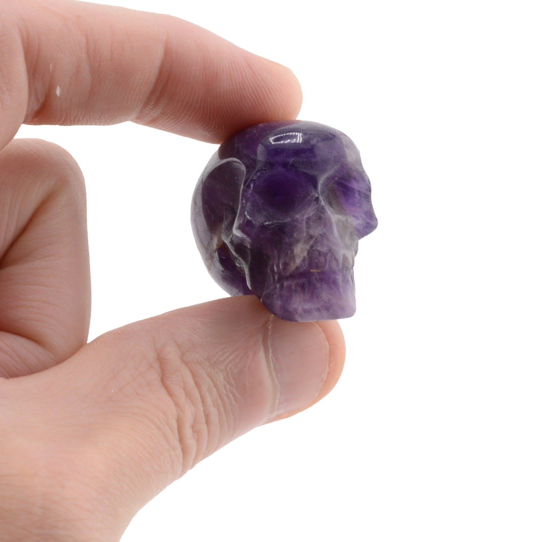 A hand holding a small carved purple amethyst skull against a plain white background.