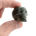 A hand holding a small carved green moss agate gemstone skull against a plain white background.