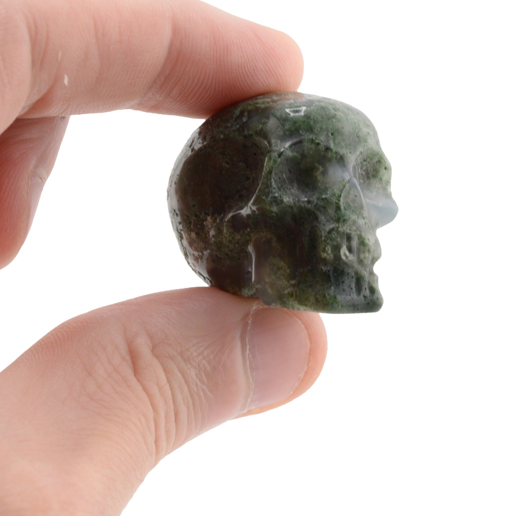 A hand holding a small carved green moss agate gemstone skull against a plain white background.
