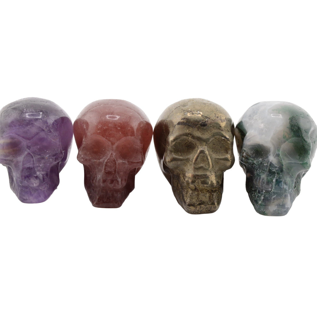 Four polished crystal skull carvings in purple, reddish-brown, metallic gold, and green marbled stone (amethyst,pyrite, moss agate & raspberry aventurine), arranged in a row on a plain white background.