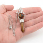 A tiger's eye crystal point and merkaba pendulum, held in a person's open hand against a white background.
